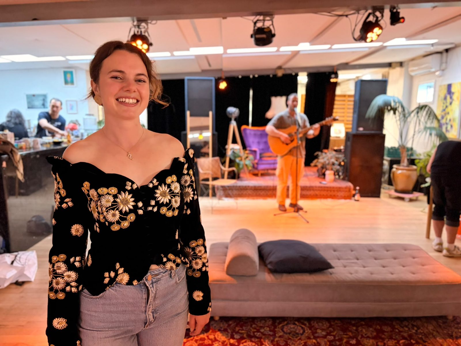 Kate smiling at her book launch in a black top embroidered with sunflowers; behind her Carl o Yāng plays guitar on the small stage.