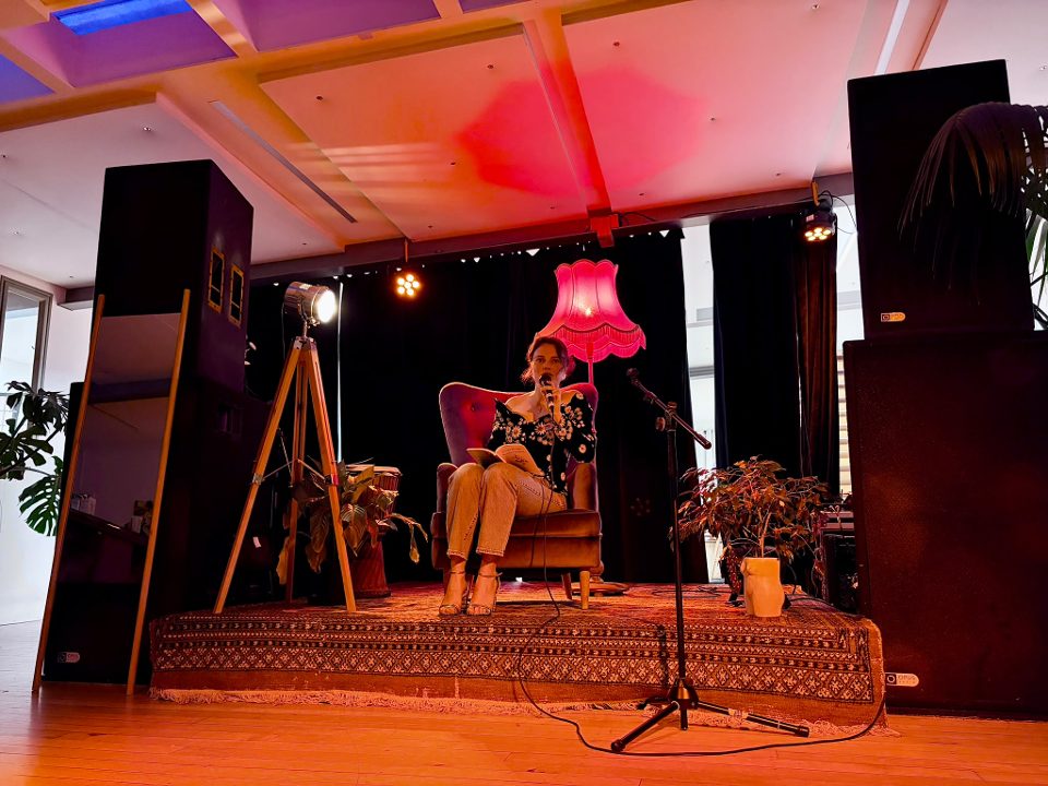 Kate reading on stage from an armchair, under a pink fringed lamp.