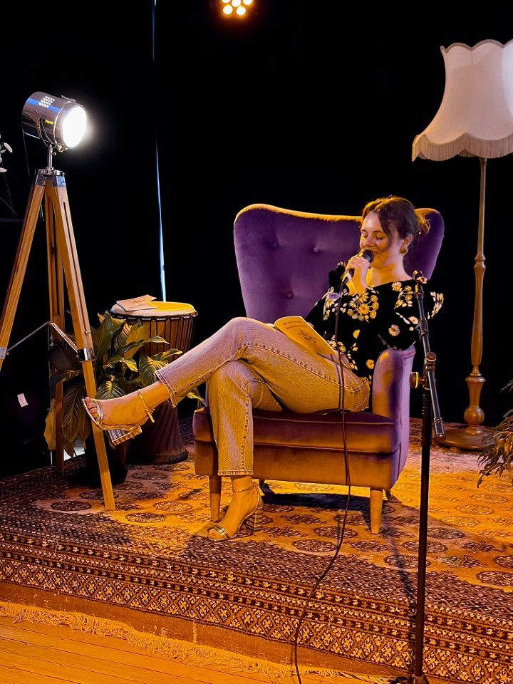 Kate reading from the book into a microphone, legs crossed in a purple armchair.